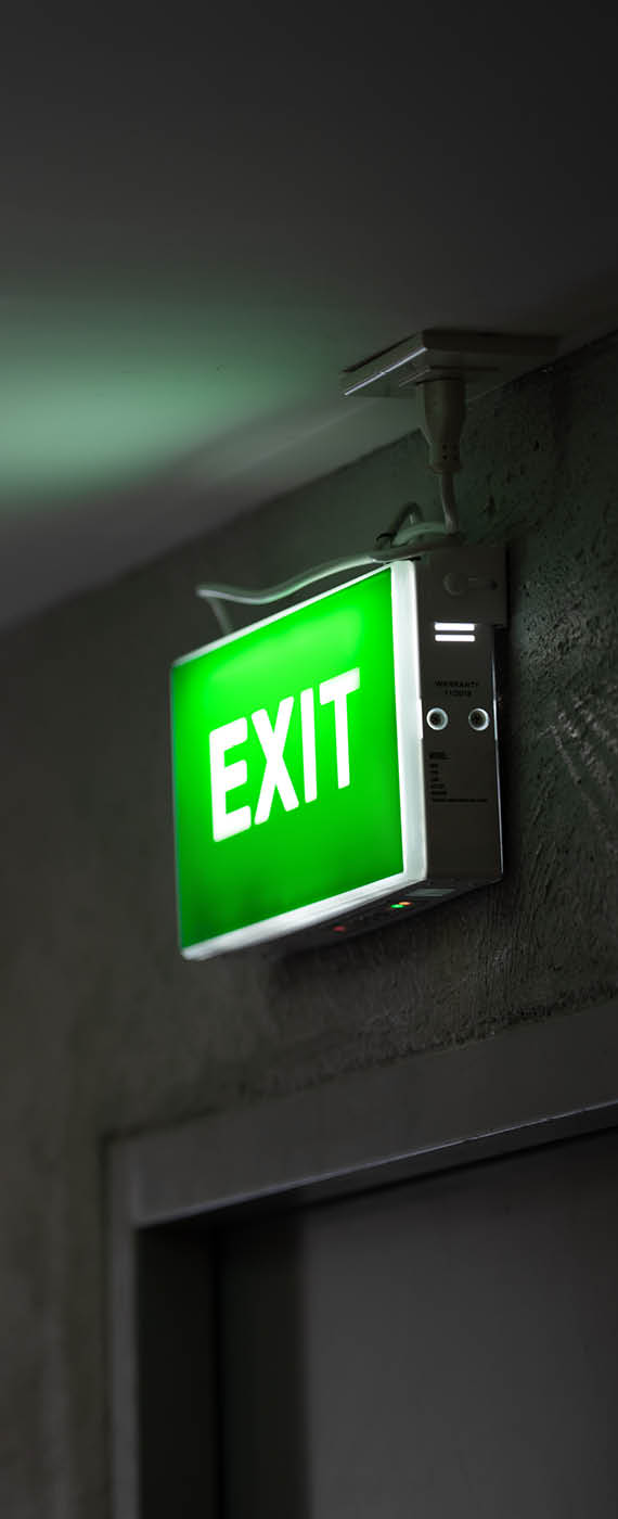 A green fire exit sign is placed on the ceiling along the dimly lit corridor and there is green exit sign on the exit door.